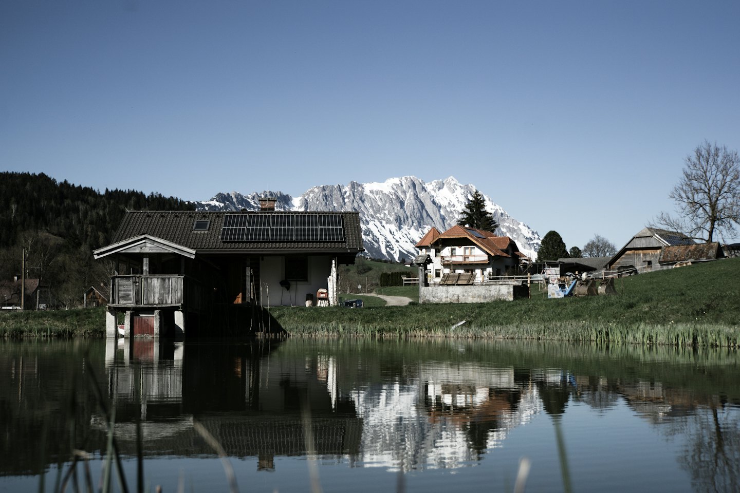 Boathouse and houses reflected in a lake below snowy mountains.